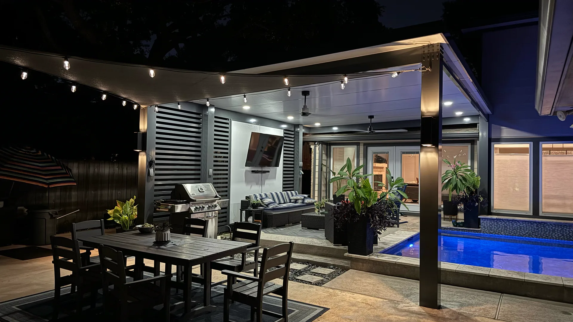 Black aluminum pergola attached to a Houston home at night, with lights on over the dining and lounge area by the pool.