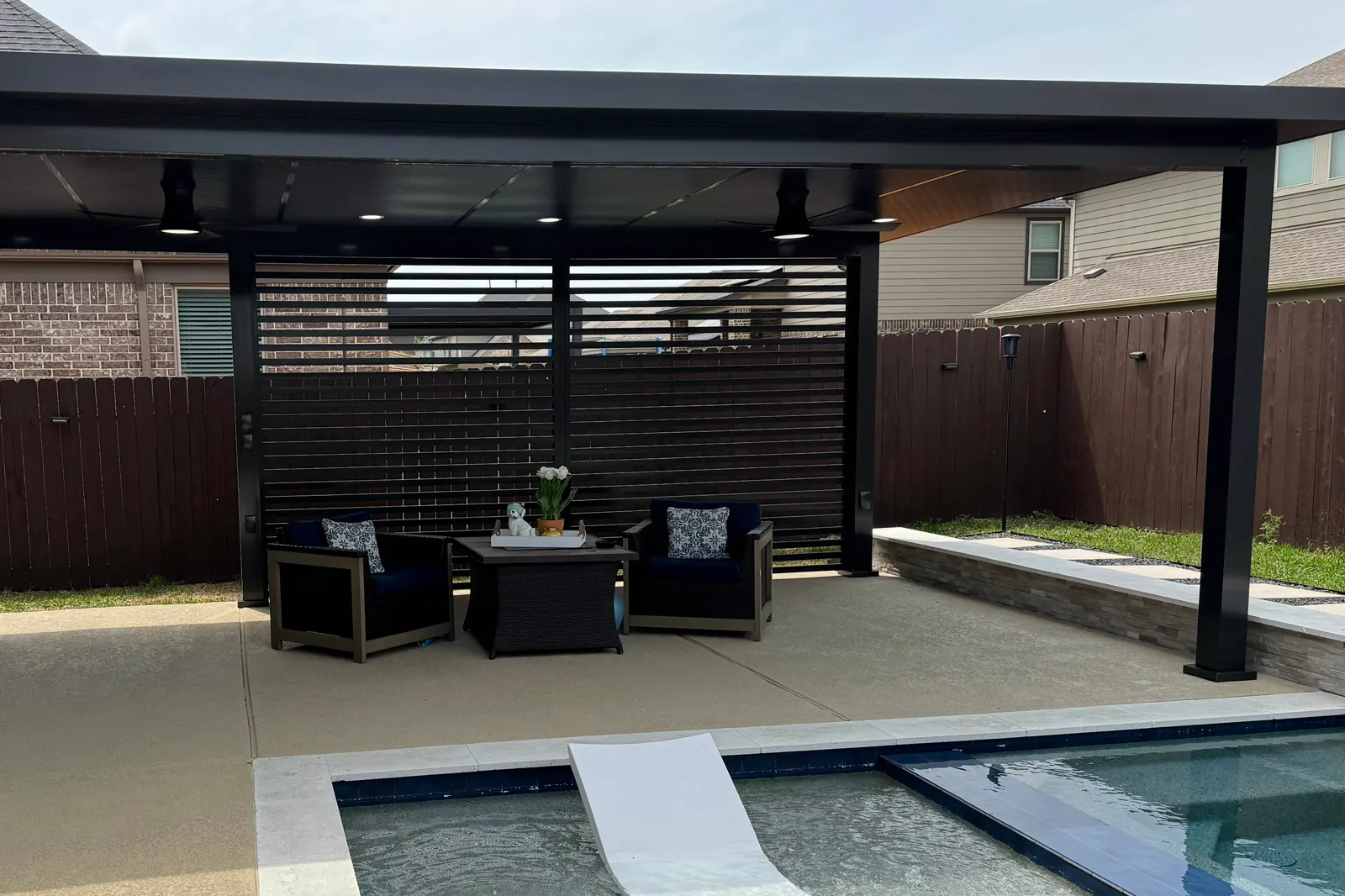 Sillones de exterior con cojines bajo un techo de aluminio negro con celosías de privacidad, junto a una piscina.